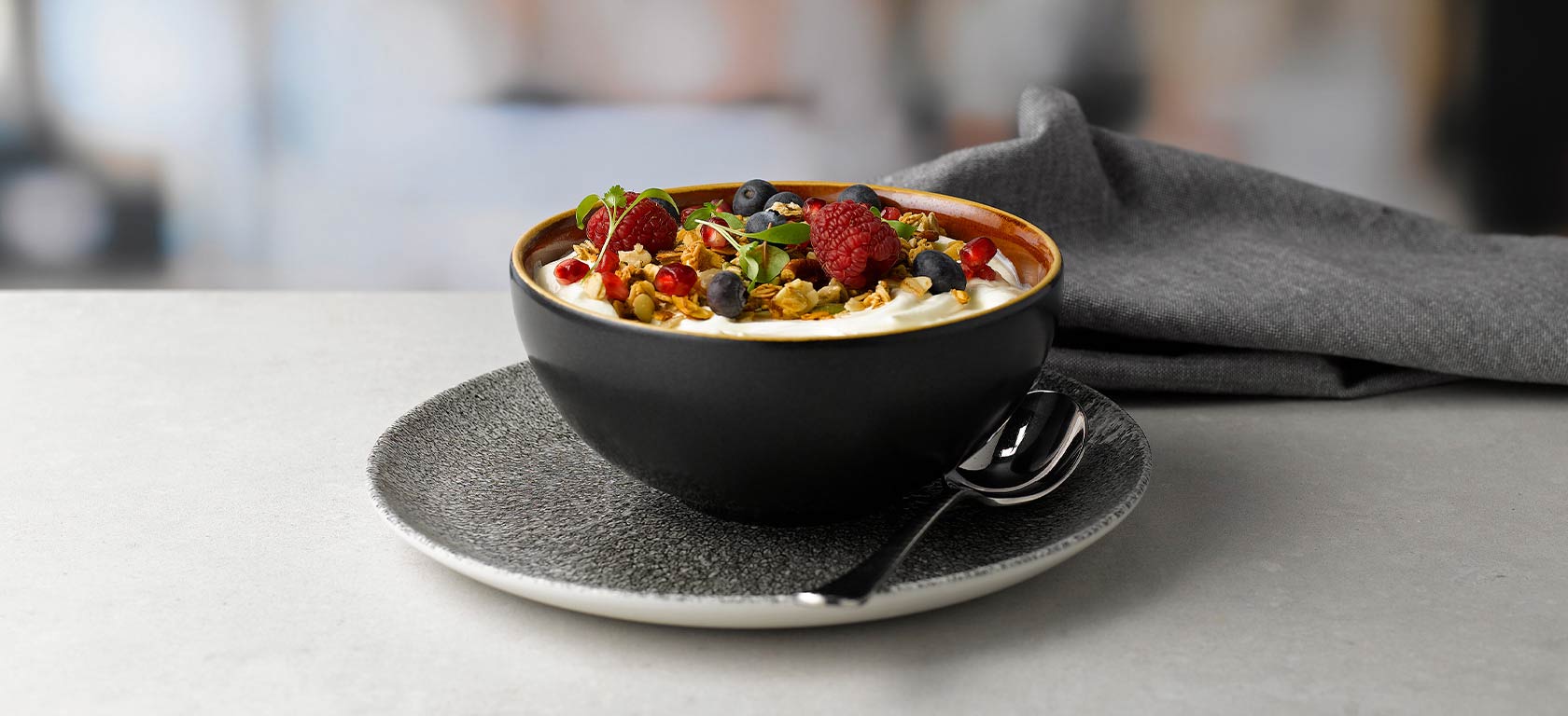 Black bowl sitting on grey saucer containing yogurt and fruit.