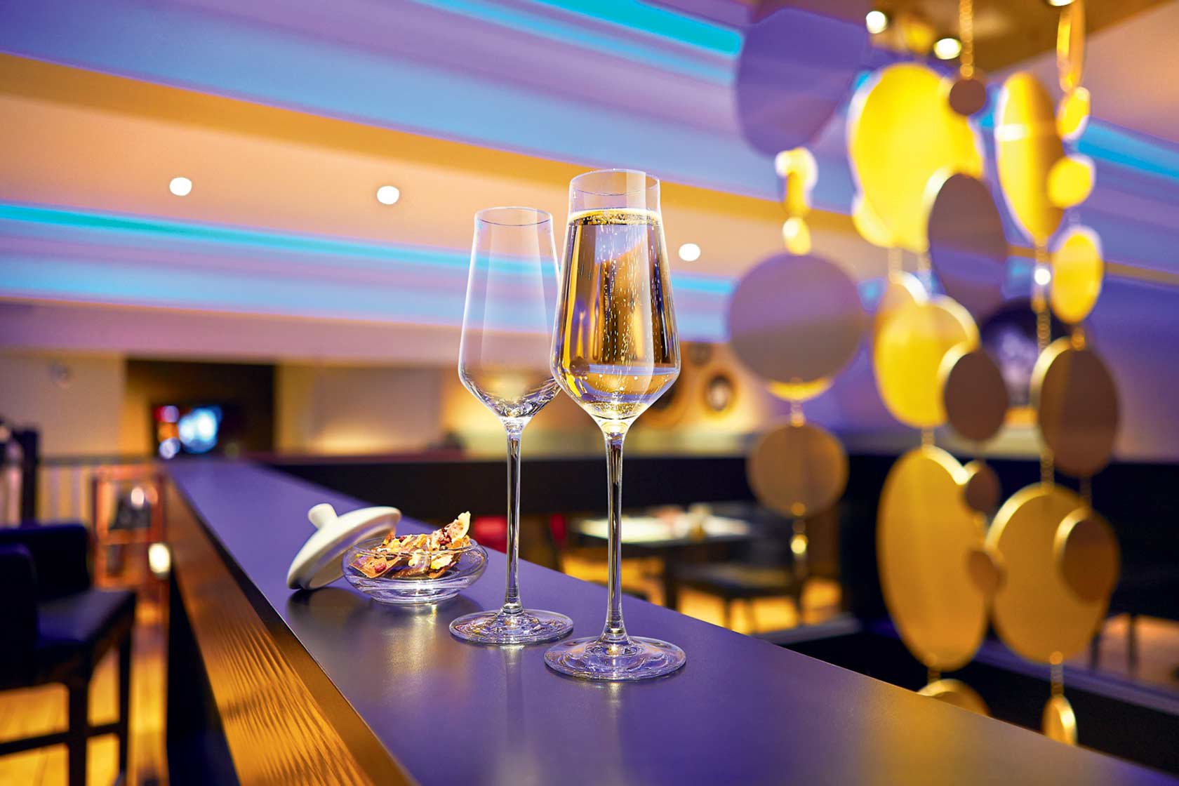 Upscale glass champagne flutes in the foreground, festive low-light decorations in the background.